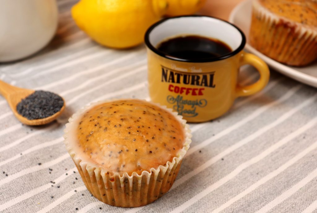 foto mostra muffins de limão com semente de papoula a frente e cenário com xícara de café, limões, prato com muffins e uma colher com semente de papoula